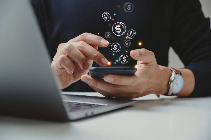 A man is sitting at a desk with an open laptop as he types away on his smartphone and dollar signs come out of the screen