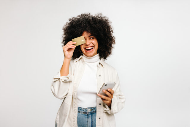 Cheerful young woman holding a credit card and a smartphone against a white background.