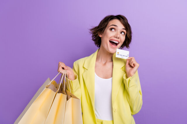 Cheerful woman carrying shopping bags and credit card against a purple background.