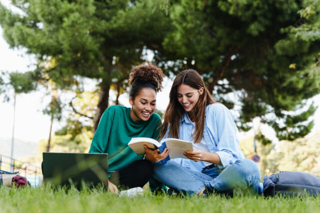 Two young female college students sitting in the grass and reading a book.