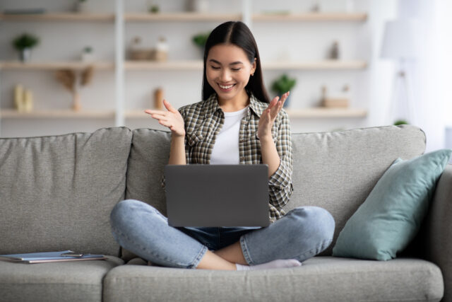 Young Asian woman smiles happily at her laptop while sitting at home on her couch.