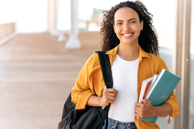 Cheerful Hispanic woman smiles while holding a bookbag and textbooks.