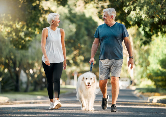 Senior couple walking dog in nature smile happily at one another.