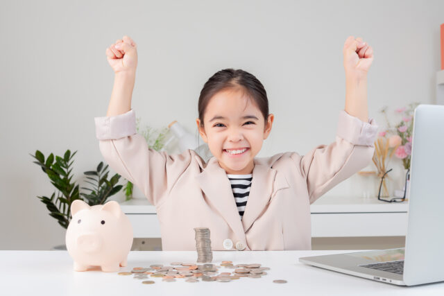 Little Asian girl in cream suit smiles triumphantly at table with piggy bank and coins.