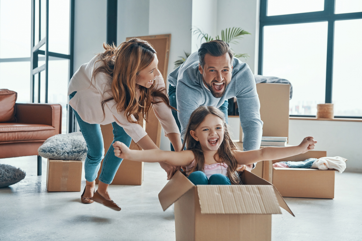 Cheerful young family moving into new home and pushing around daughter in moving box.