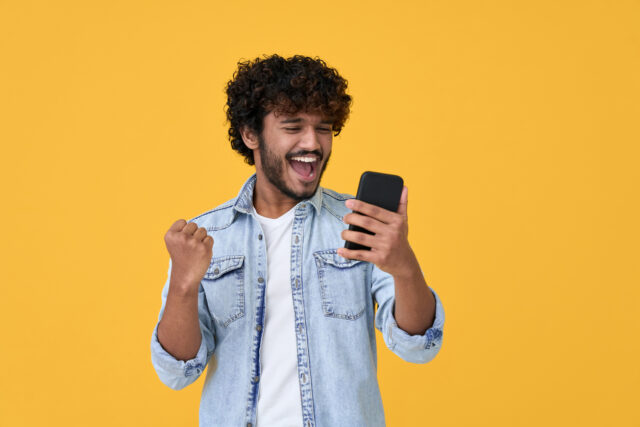 Excited young Indian man pumps fist in air while looking at smart phone against yellow background.
