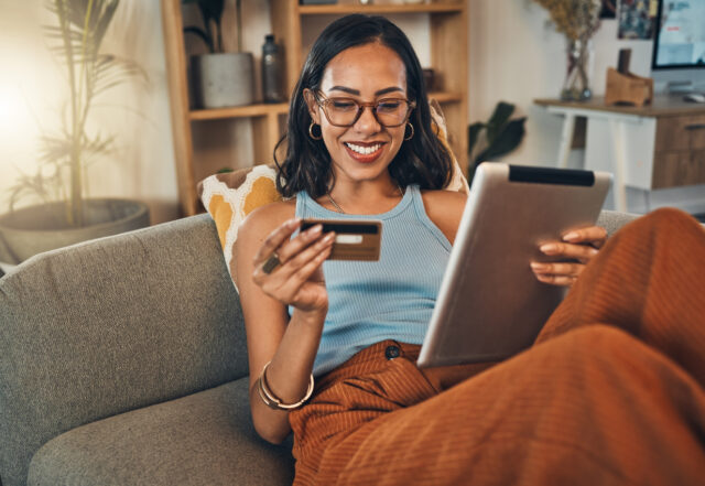 Smiling mixed race woman sitting on sofa smiles while using tablet and holding credit card.