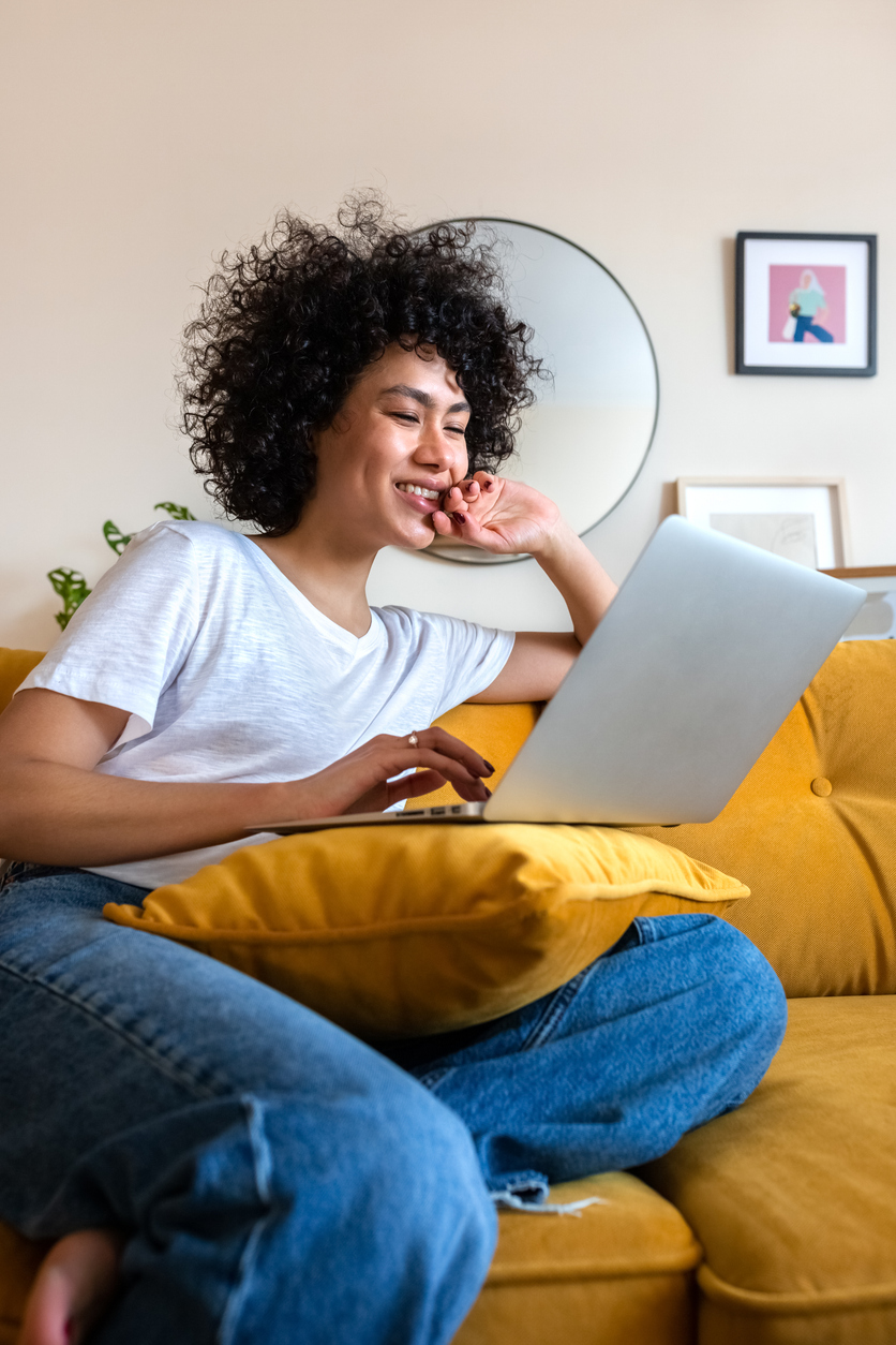 Happy African American woman with afro contentedly using laptop while sitting on yellow sofa.