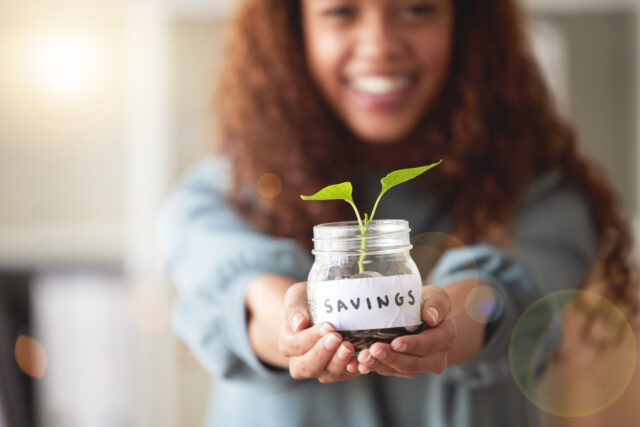 Young African American woman holds hands out with savings jar where a small plant grows.