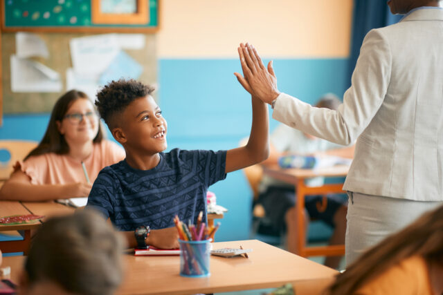 Happy African American boy high fives teacher in classroom.
