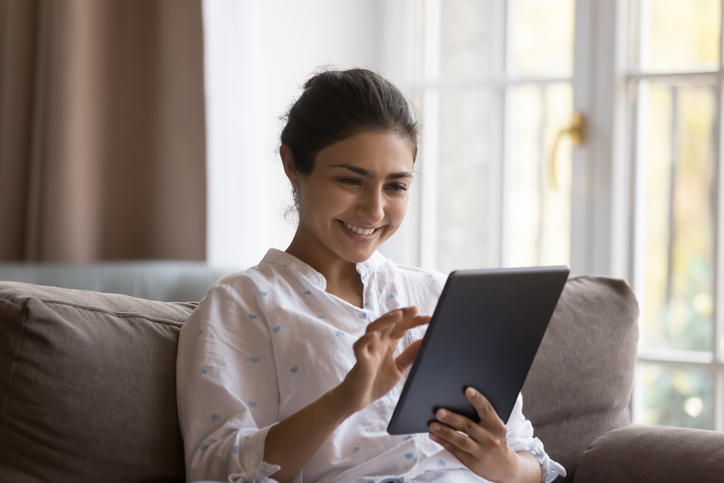 Woman sitting on couch smiles as she reads on smart tablet.