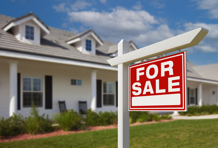 House sits on street during a sunny day with a For Sale sign on its front lawn. 