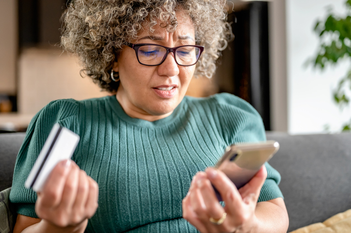 Woman stares frustratedly at her phone in one hand and her debit card in another