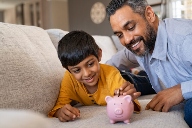 Indian father watches his young son put coins in a piggy bank while sitting on couch.
