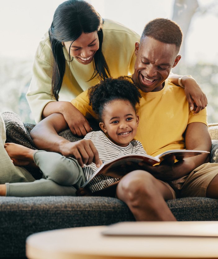 Happy African American family sitting on couch reading book to child.