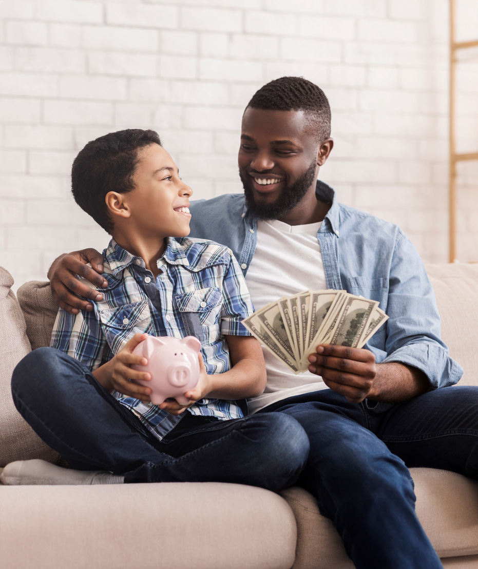 Child sits with father on couch with piggy bank and money in hands.