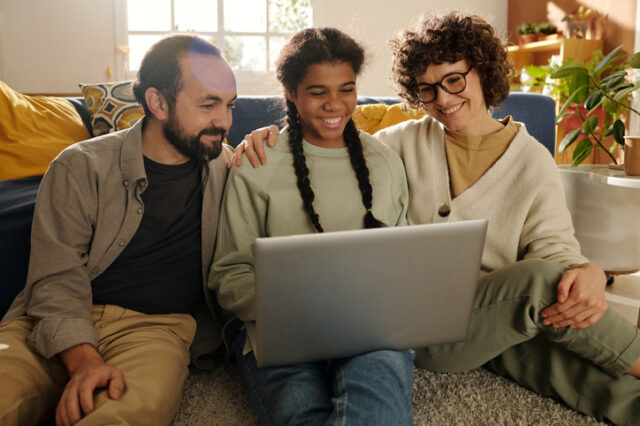 Young teen girl sits on floor in between parents as they all happily look at the laptop on her lap.
