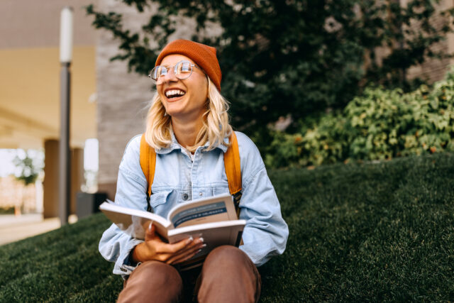 Young blonde woman with orange hat smiles on college campus while reading book.