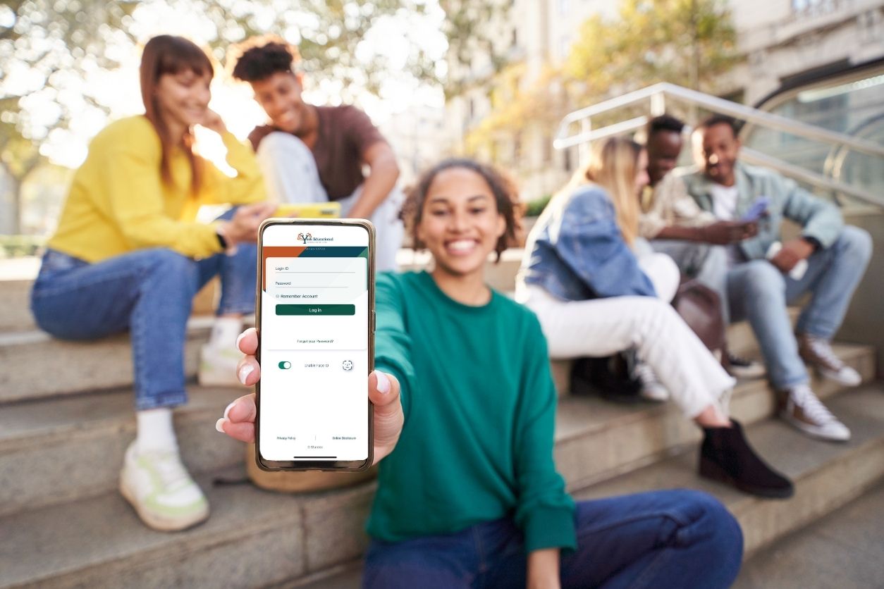 Students sit outside on stairs while one holds up her phone with the YEFCU mobile app open on it.