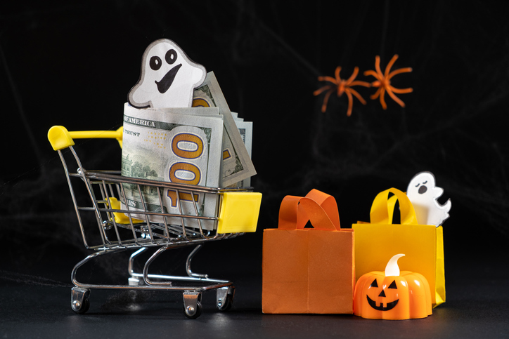 On a black background, a miniature grocery cart is filled with hundred dollar bills and the paper cut out of a ghost. It's surrounded by small paper cut outs of shopping bags and pumpkins.