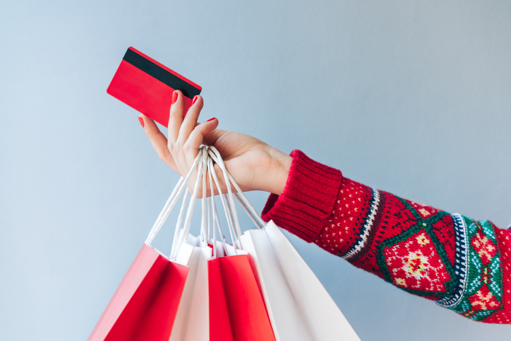 A woman's arm wearing christmas sweater holds a red credit card in her hand alongside multiple shopping bags.