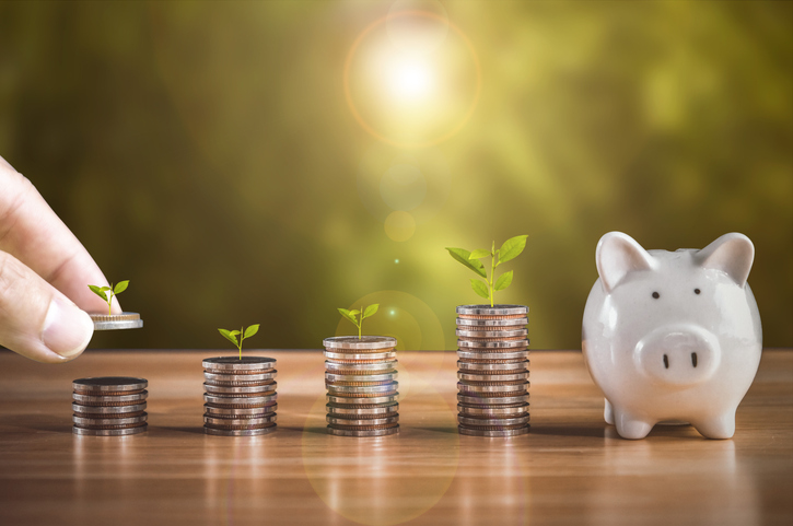 Growing stacks of coins sit on a table leading up to a piggy bank, implying the saving of money.