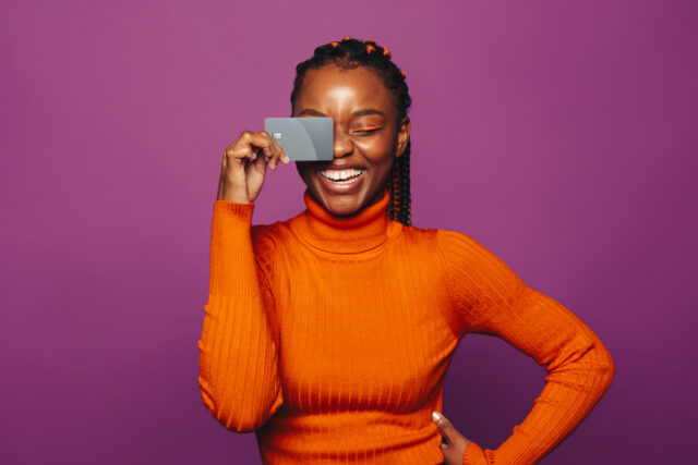 Young African American woman in orange sweater smiles as she holds a credit card up near her face.