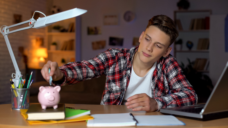Teen boy sitting at his at home desk puts cash into a piggy bank.